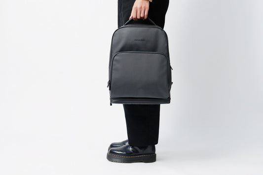 Person in black trousers and boots holding the Fetchamo Core Backpack by its top handle, matte black finish — featured image.
