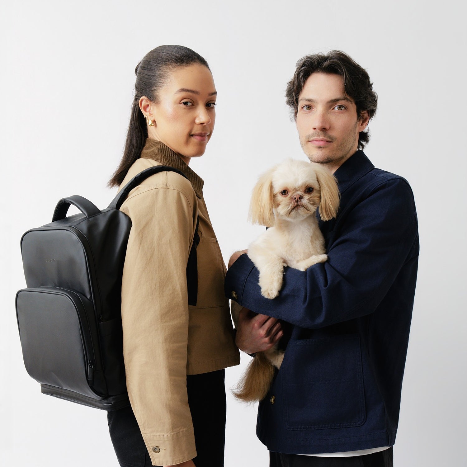 Founders of Fetchamo posing with their small white Shih Tzu, with the black Fetchamo Core Backpack, against a light grey studio background.