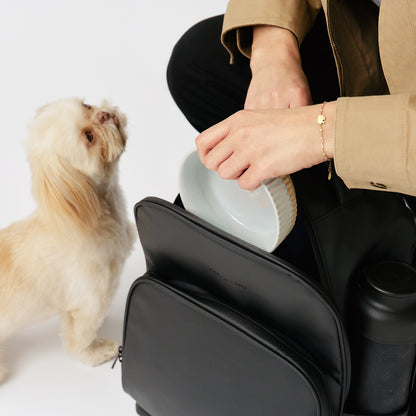 fetchamo Core backpack for dog owners – model placing a dog bowl into the backpack with dog beside her