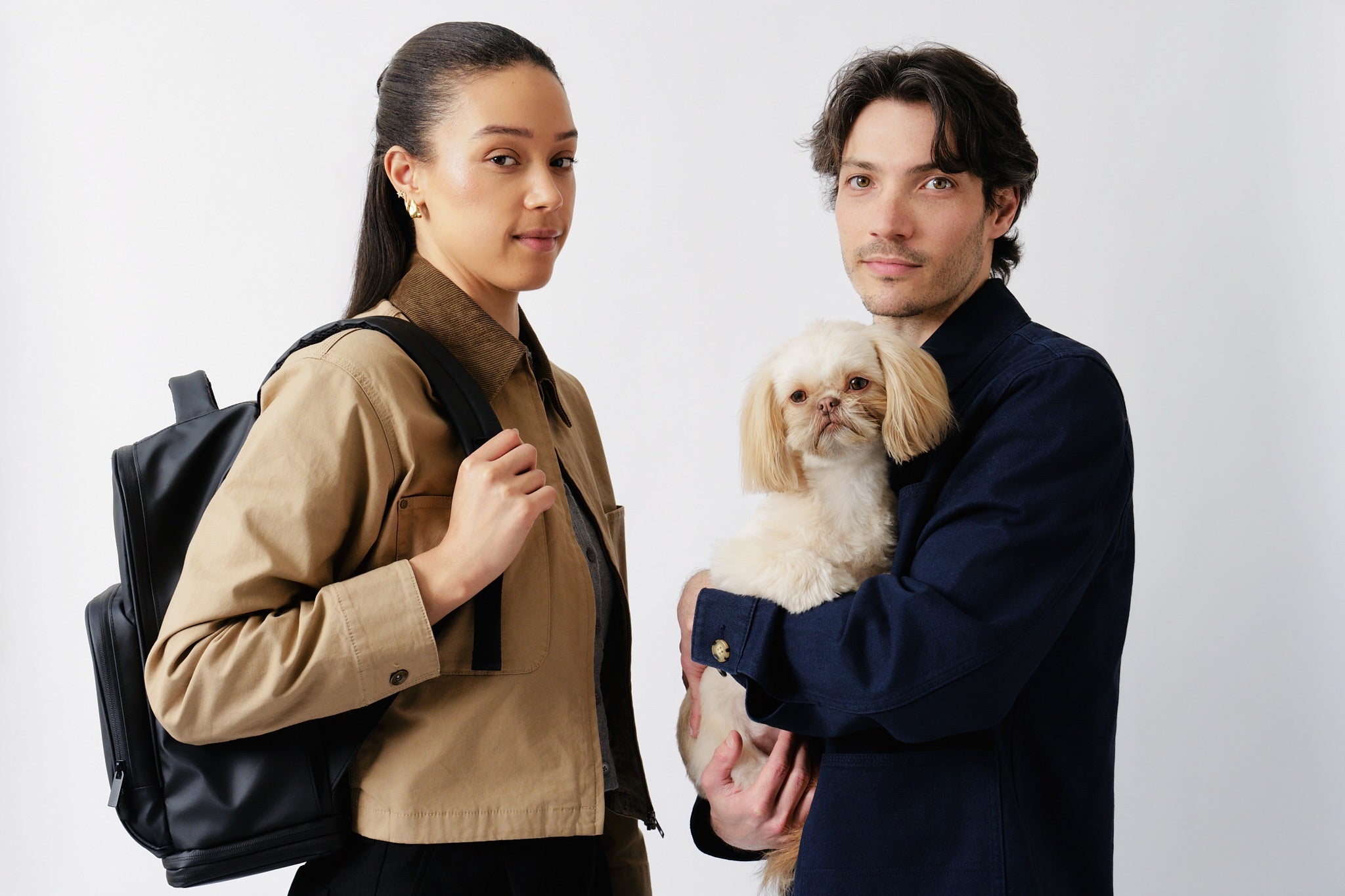 Founders of Fetchamo posing with their small white Shih Tzu, with the black Fetchamo Core Backpack, against a light grey studio background.